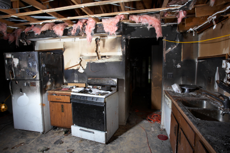 A heavily damaged kitchen with soot-covered appliances and exposed ceiling materials shows the aftermath of a residential fire.
